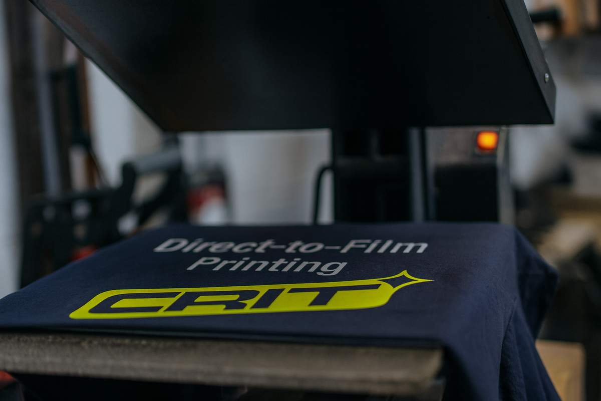 Direct-to-Film (DTF) printing process at Crit Apparel, applying neon yellow CRIT logo to a navy U.S.-made t-shirt using a heat press